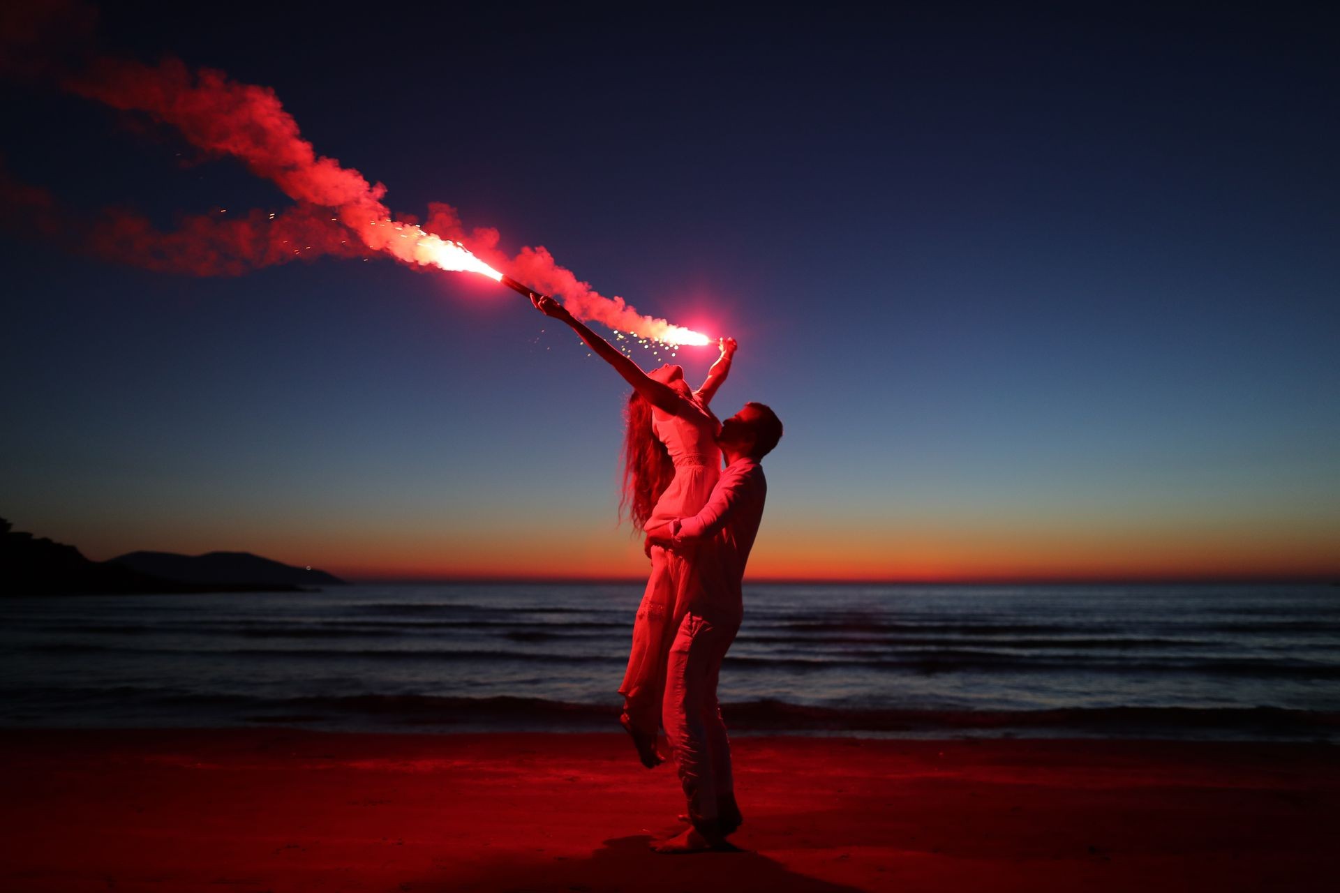 Couple on beach under night sky, holding a red flare with ocean in background.